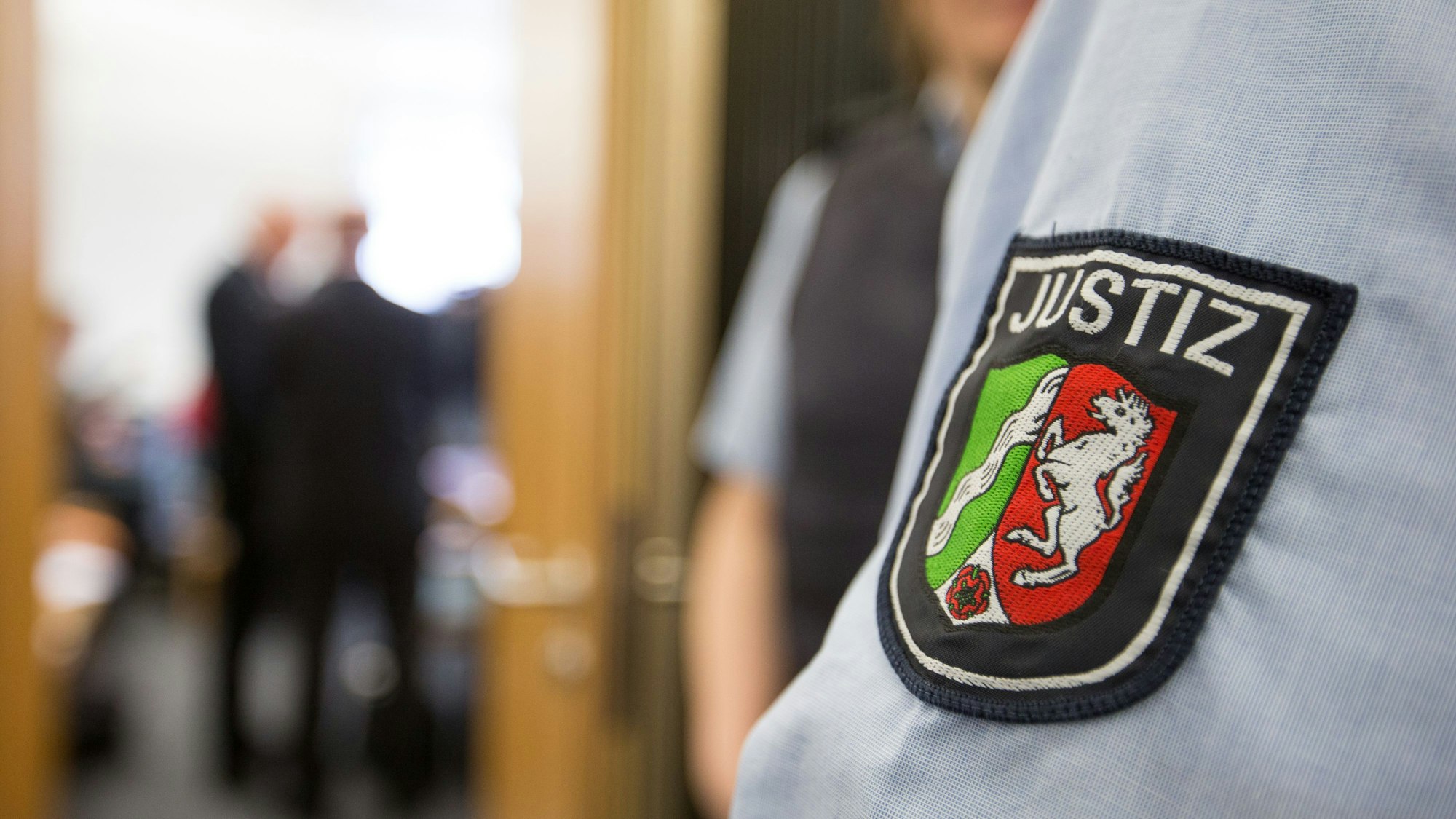 Ein Justizbeamter steht beim Prozessauftakt vor dem Sitzungssaal. Nur sein Ärmel mit dem NRW-Wappen ist zu sehen.