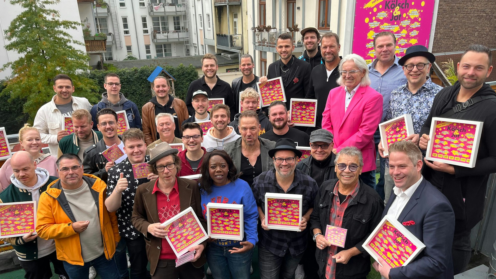 Kölner Künstler stehen in einem Pulk auf einer Terrasse, einige halten einen Rahmen mit dem pinken Cover der CD „Kölsch & Jot“.
