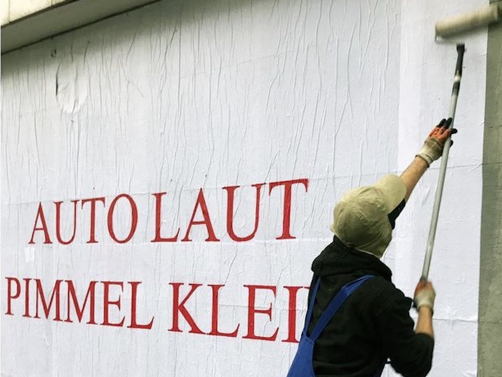 Mann im Blaumann, der mit einer Farbrolle und einem Eimer ein Plakat an einer Wand anbringt