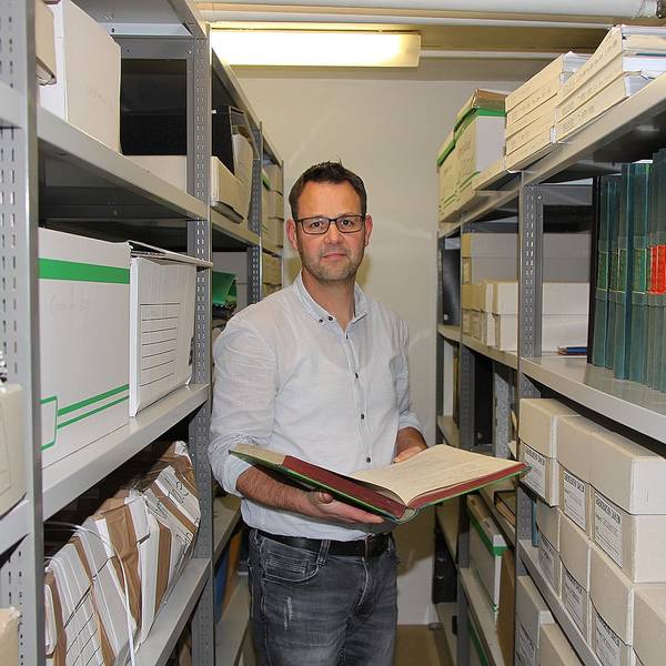 Der Dahlemer Kämmerer Frank Hütter steht zwischen zwei hohen Regalen im Gemeindearchiv. Er hält eine aufgeschlagene Akte in der Hand.
