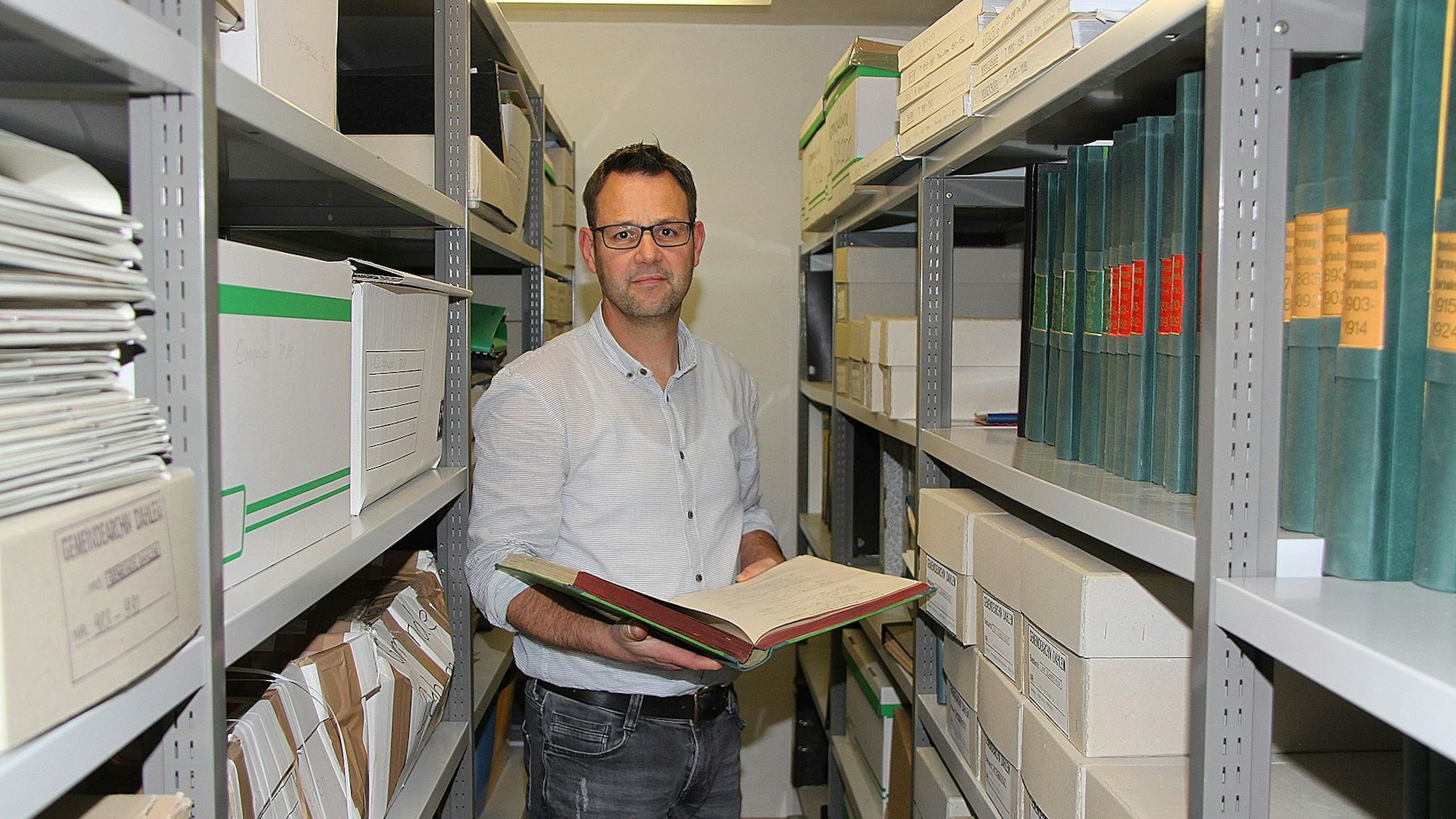 Der Dahlemer Kämmerer Frank Hütter steht zwischen zwei hohen Regalen im Gemeindearchiv. Er hält eine aufgeschlagene Akte in der Hand.