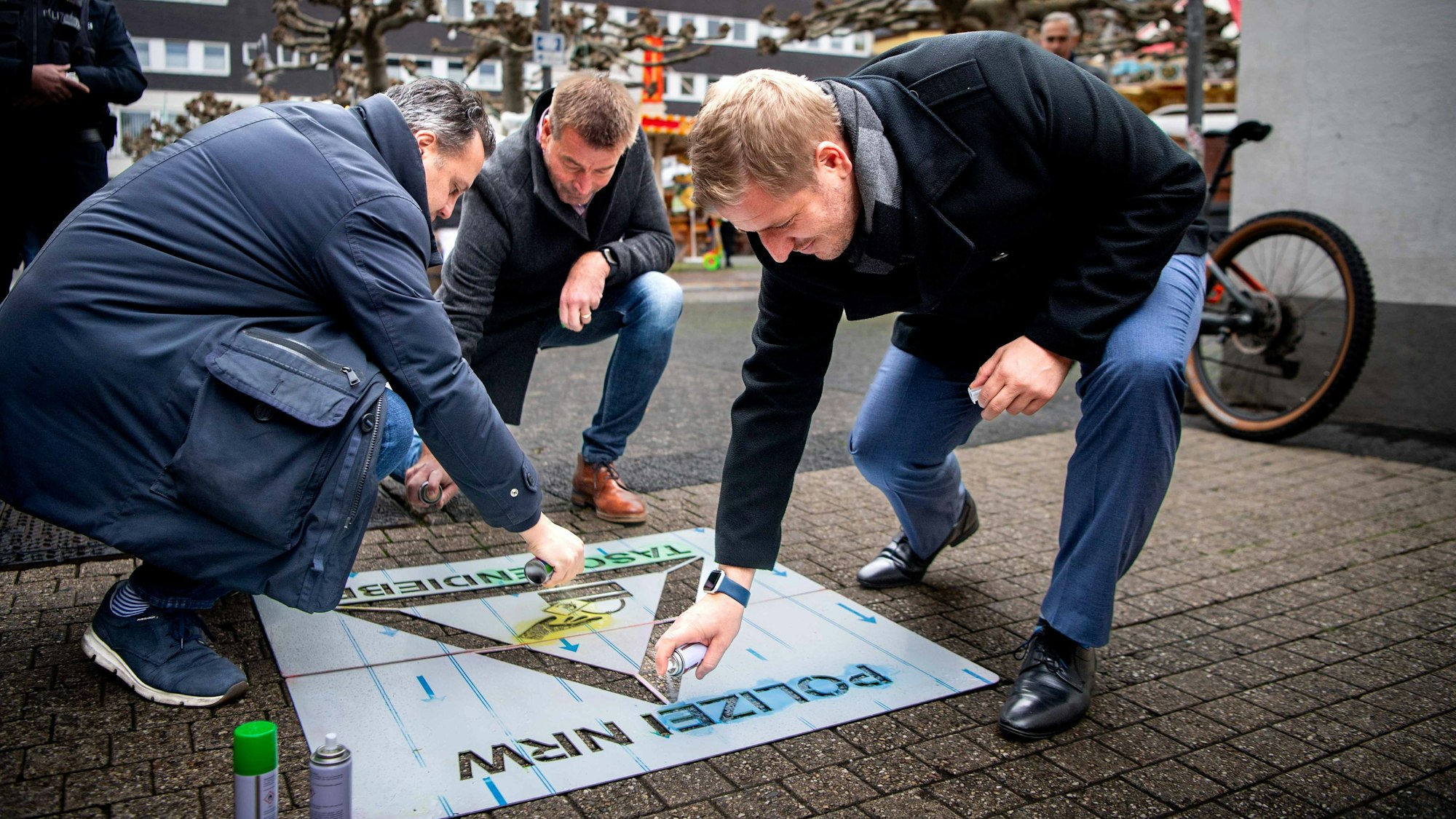 Das Bild zeigt Sacha Reichelt, Ulrich Linden und Markus Ramers, wie sie mit einer Sprühflasche ein Piktogramm mithilfe einer Schablone auf den Boden aufbringen.