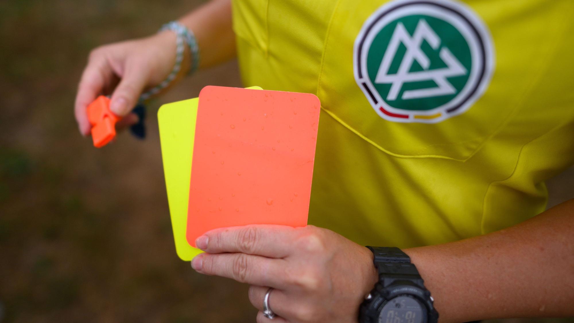 Eine Schiedsrichterin steht mit roter und gelber Karte auf einem Rasen und hält in der anderen Hand eine Trillerpfeife.