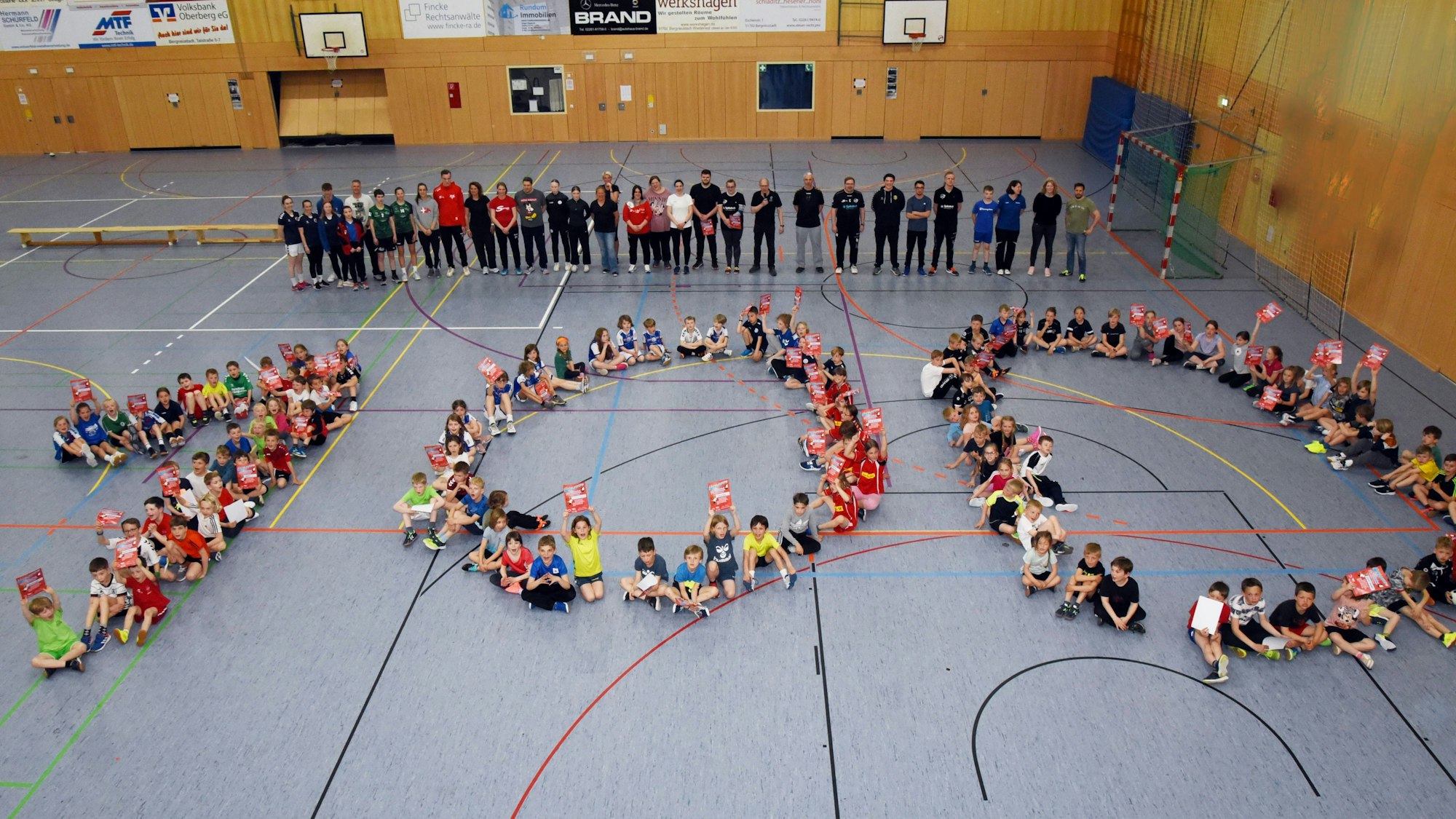 Zahlreiche Kinder bilden auf einem Hallenboden die Zahl 100.