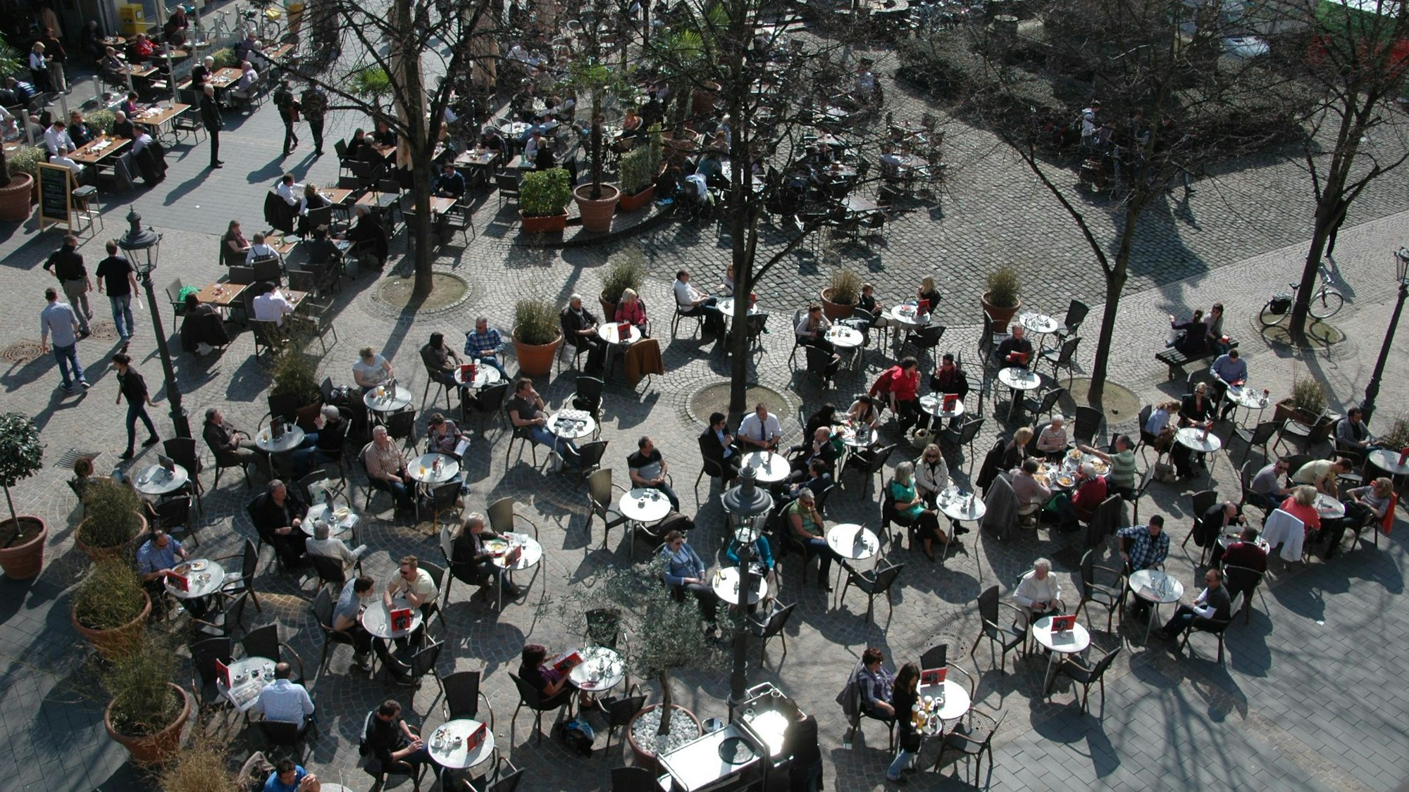 Gäste sitzen an den Tischen eines Gastronomischen Betriebes auf einem Platz.