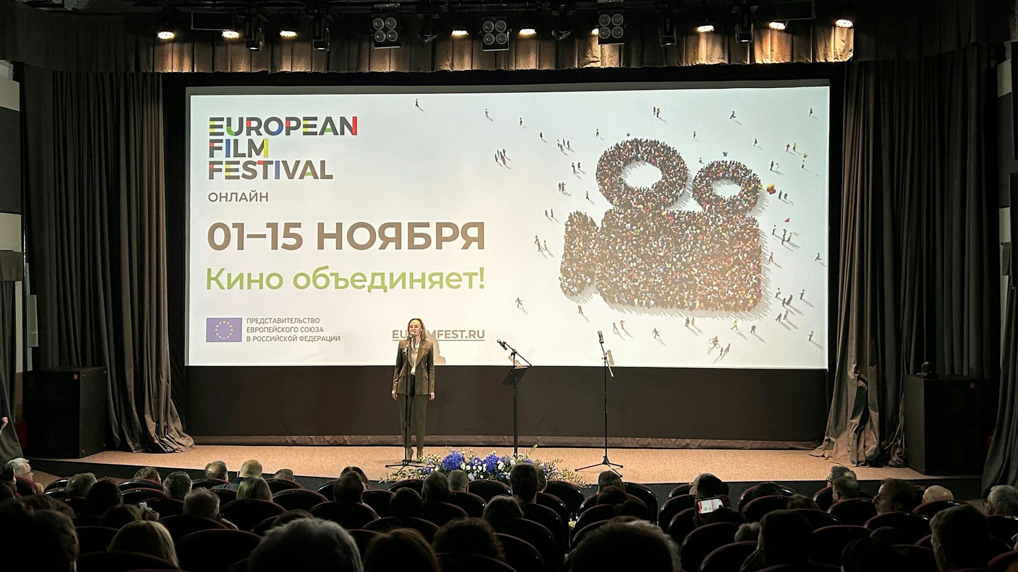 Die Leinwand in einem Kinosaal zeigt eine Menschenmasse auf einem weißen Hintergrund von oben, die sich in Form einer Kamera zusammengestellt hat. Auf der Bühne davor steht eine Frau an einem Mikrofon.