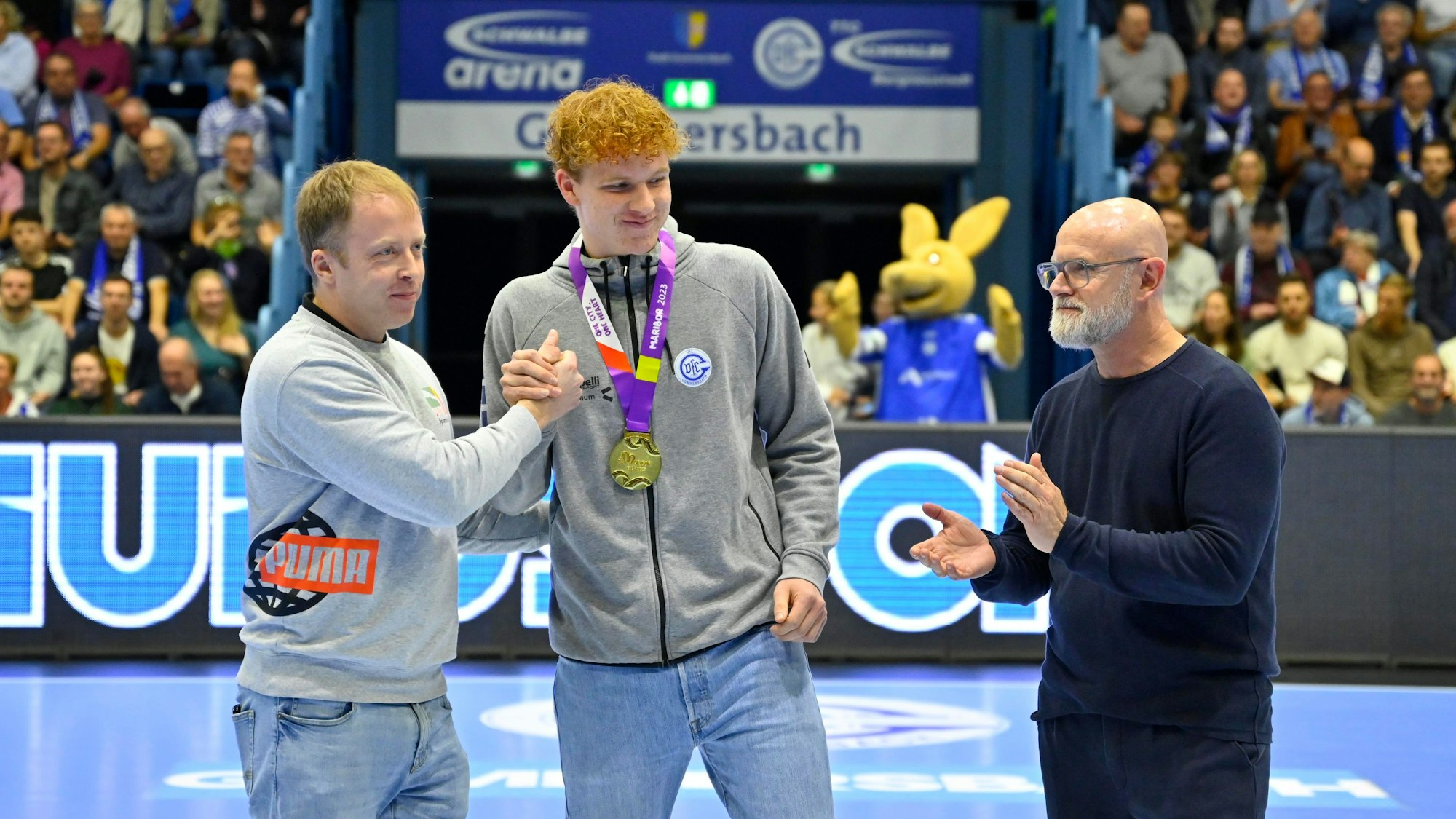 Zwei Männer gratulieren einem jungen Handballer, nachdem sie ihm eine Goldmedaille um den Hals gehängt haben.