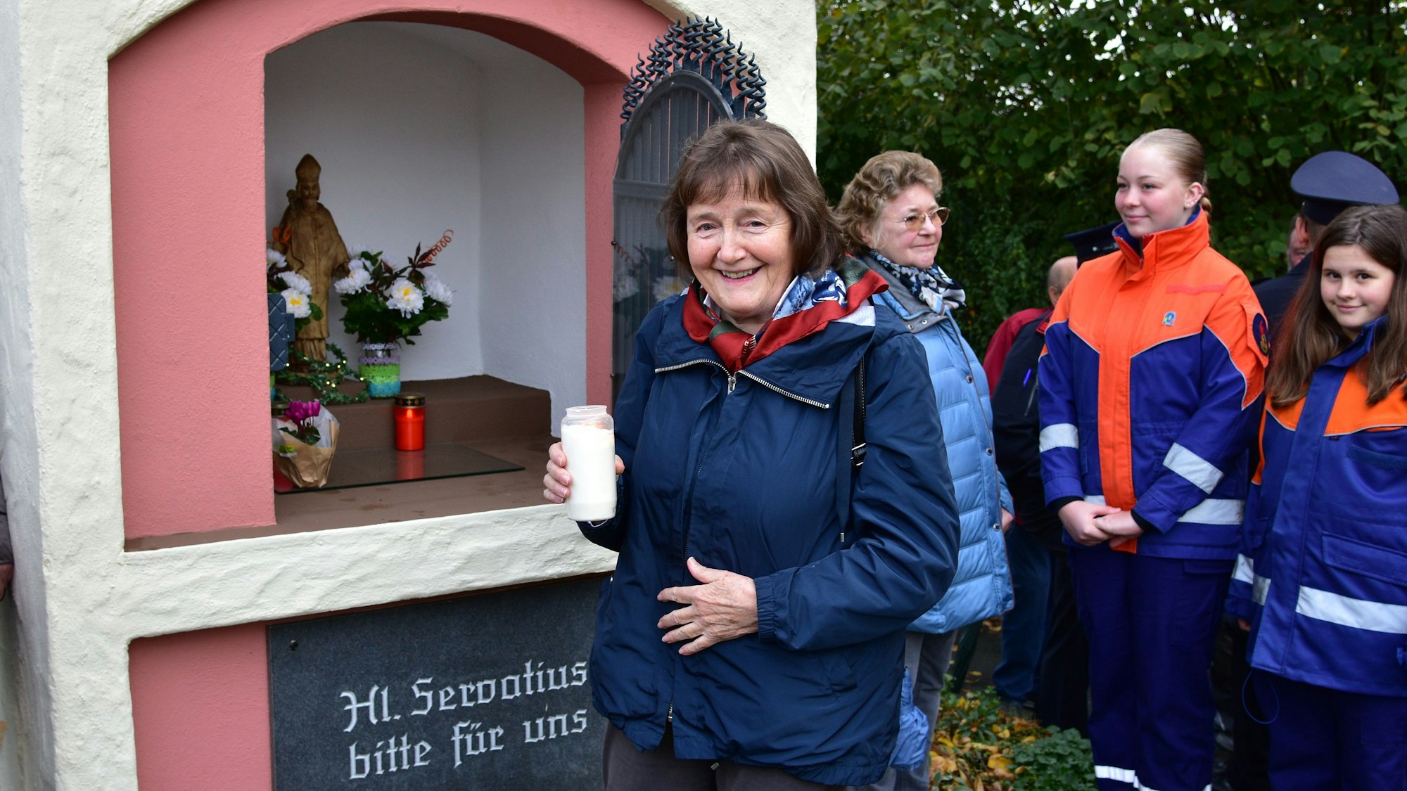 Frau hält Kerze in linker Hand, hinter ihr ein kleines Häuschen mit Blumengestecken und Kerzen