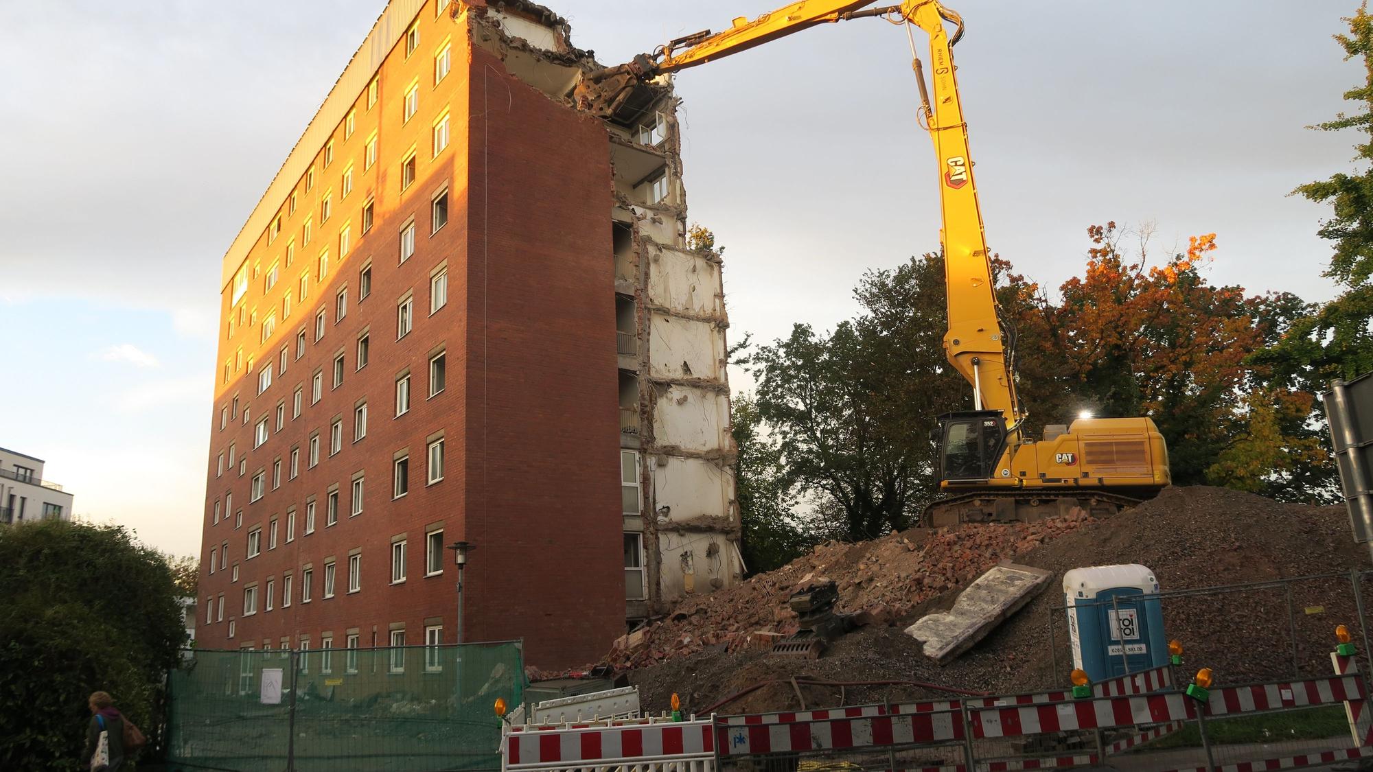 Ein Bagger, der auf einem Erdhügel steht hat seine Schaufel in die oberste Etage eines etwa zehnstöckigen Hochhauses geschlagen.