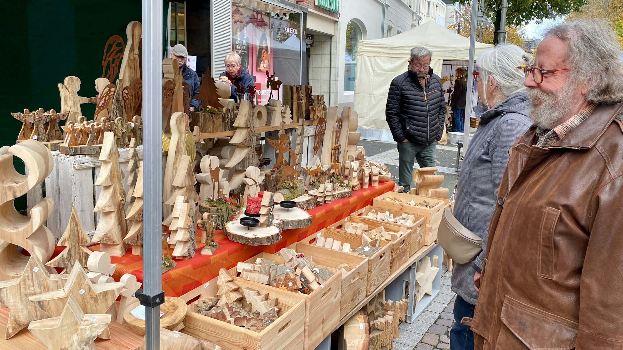 Zu sehen ist ein Verkaufsstand mit vielen Holzschnitzereien, die von Besuchern bestaunt werden.