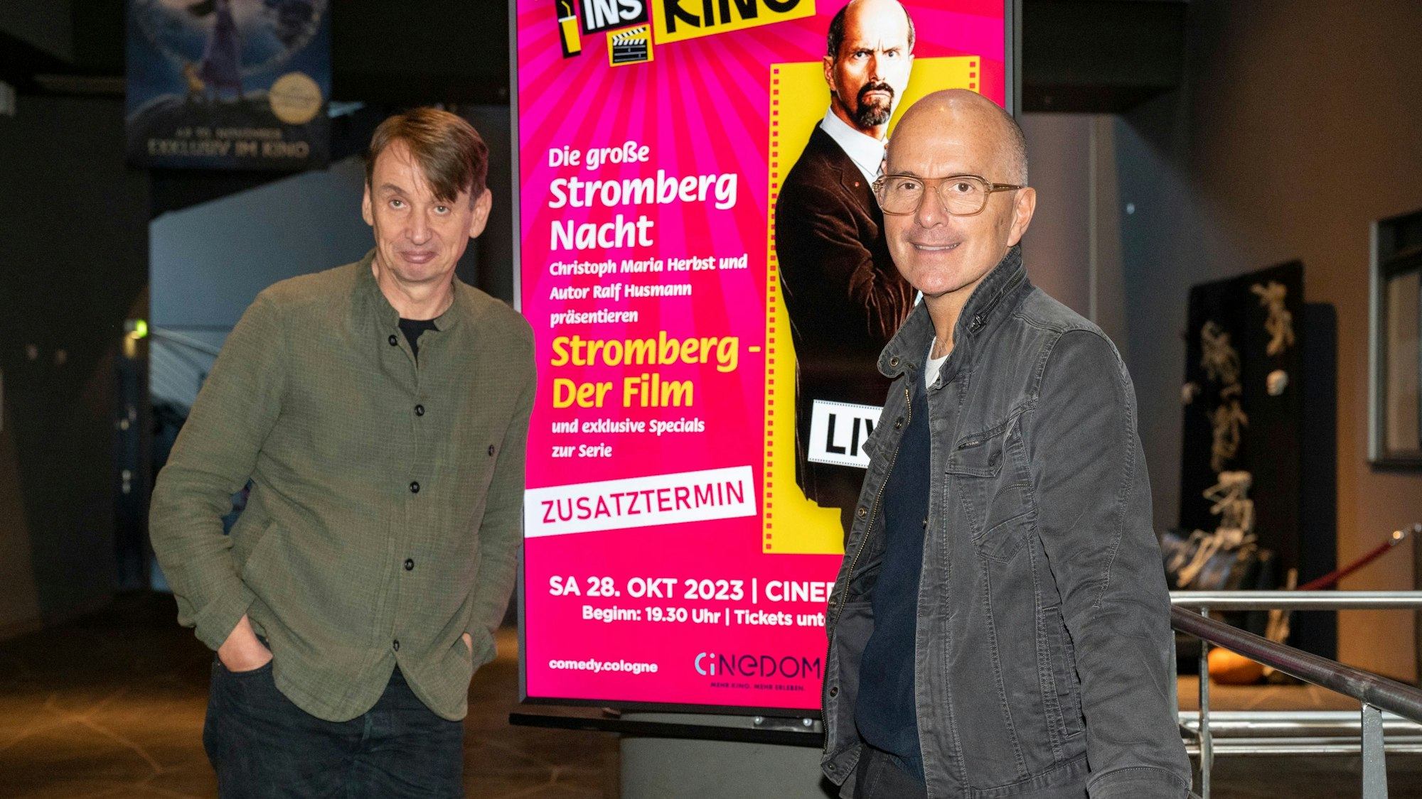 Christoph Maria Herbst und Ralf Husmann posieren neben einem digitalen Poster von Stromberg im Cinedom.