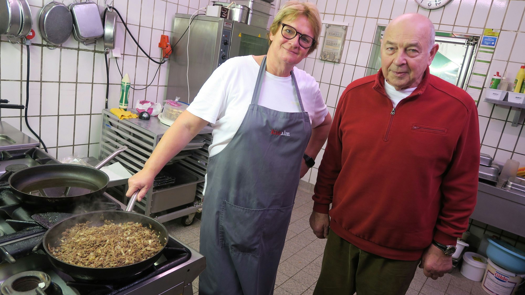 Eine Frau mit Schürze steht an einem Herd, neben ihr ein älterer Mann im roten Pullover.