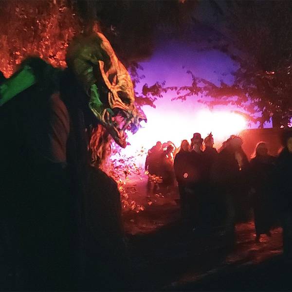 Ein Mensch mit einer gruseligen Monstermasse steht im Dunklen neben einem Weg, auf dem viele andere Menschen laufen. Die Szene ist in rot-violettes Licht getaucht.
