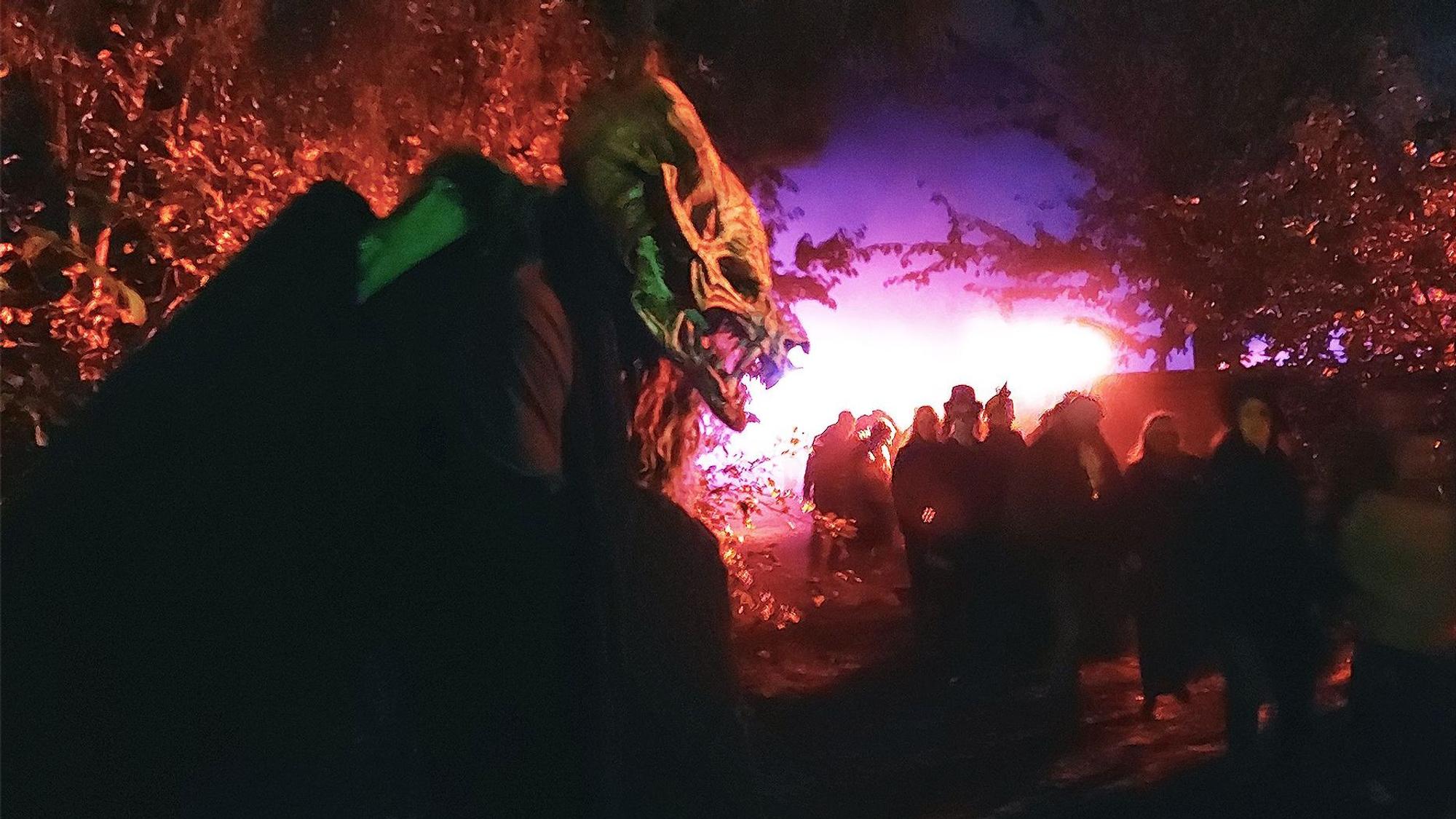 Ein Mensch mit einer gruseligen Monstermasse steht im Dunklen neben einem Weg, auf dem viele andere Menschen laufen. Die Szene ist in rot-violettes Licht getaucht.