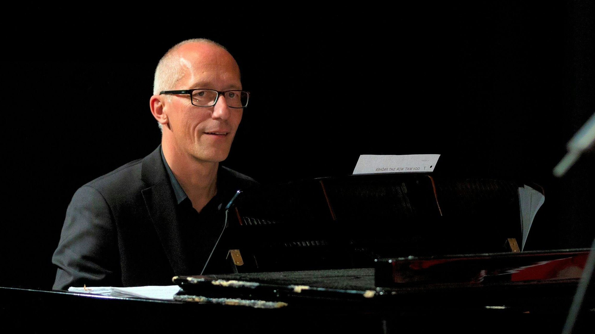 Ein Mann mit Glatze, Brille und dunklem Hemd sitzt an einem Klavier.