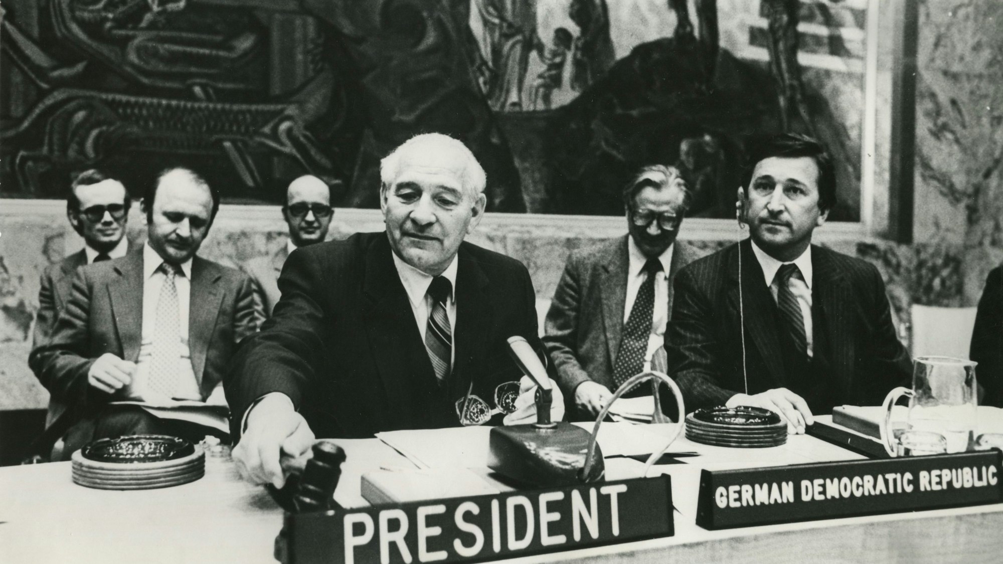 Ein älterer Mann mit dem Schild President vor sich sitzt an einem Pult. Das Bild ist in Schwarz-Weiß.