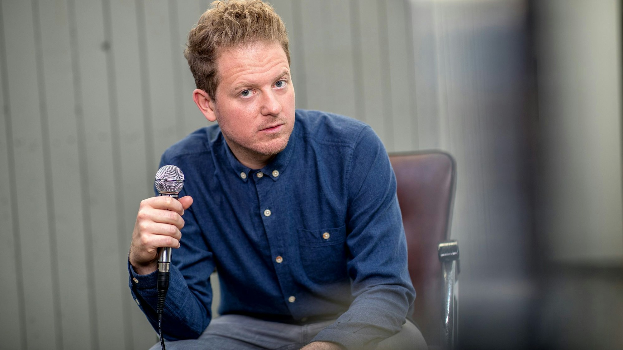 Zingsheim trägt ein blaues Hemd. Er sitzt auf einem Stuhl und hält ein Mikrofon in der Hand.