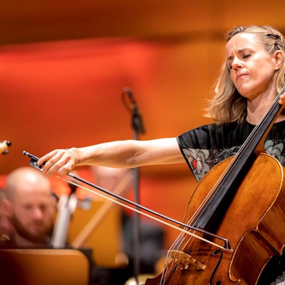 Die Cellistin gibt sich ganz ihrem Spiel hin und spielt mit geschlossenen Augen. Im Hintergrund weitere verschwommene Musiker.