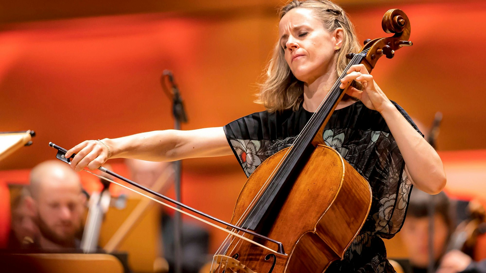 Die Cellistin gibt sich ganz ihrem Spiel hin und spielt mit geschlossenen Augen. Im Hintergrund weitere verschwommene Musiker.