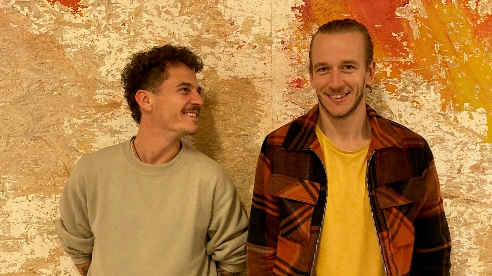 Zwei Männer stehen vor einer Wand und lächeln.