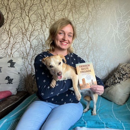 Eine Frau hat einen Hund auf dem Arm und ein hält ein Buch in der Hand.