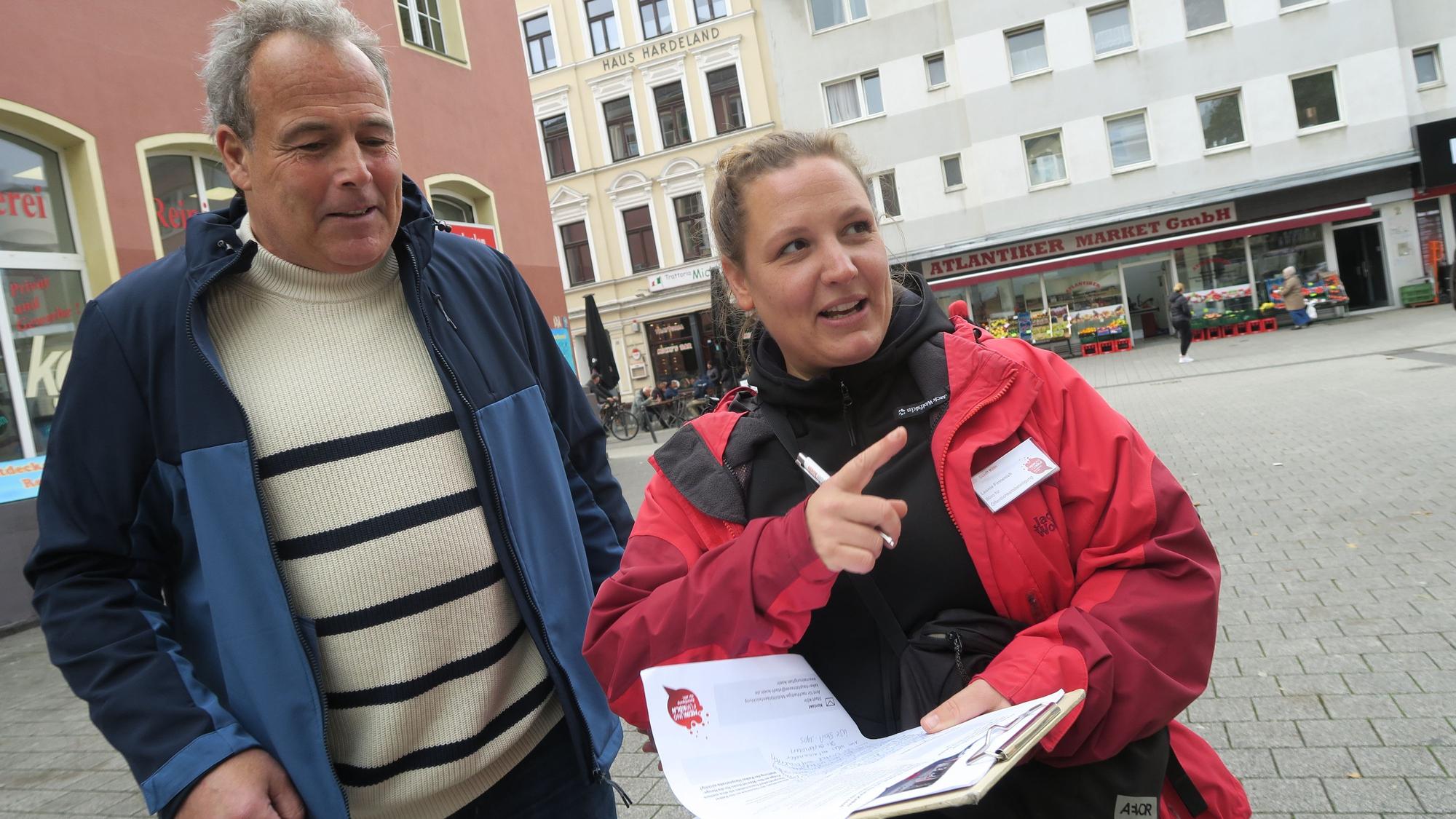 Ein Mann und eine Frau stehen auf einem Platz, die Frau hat ein Clipboard mit einem Fragebogen in der Hand.