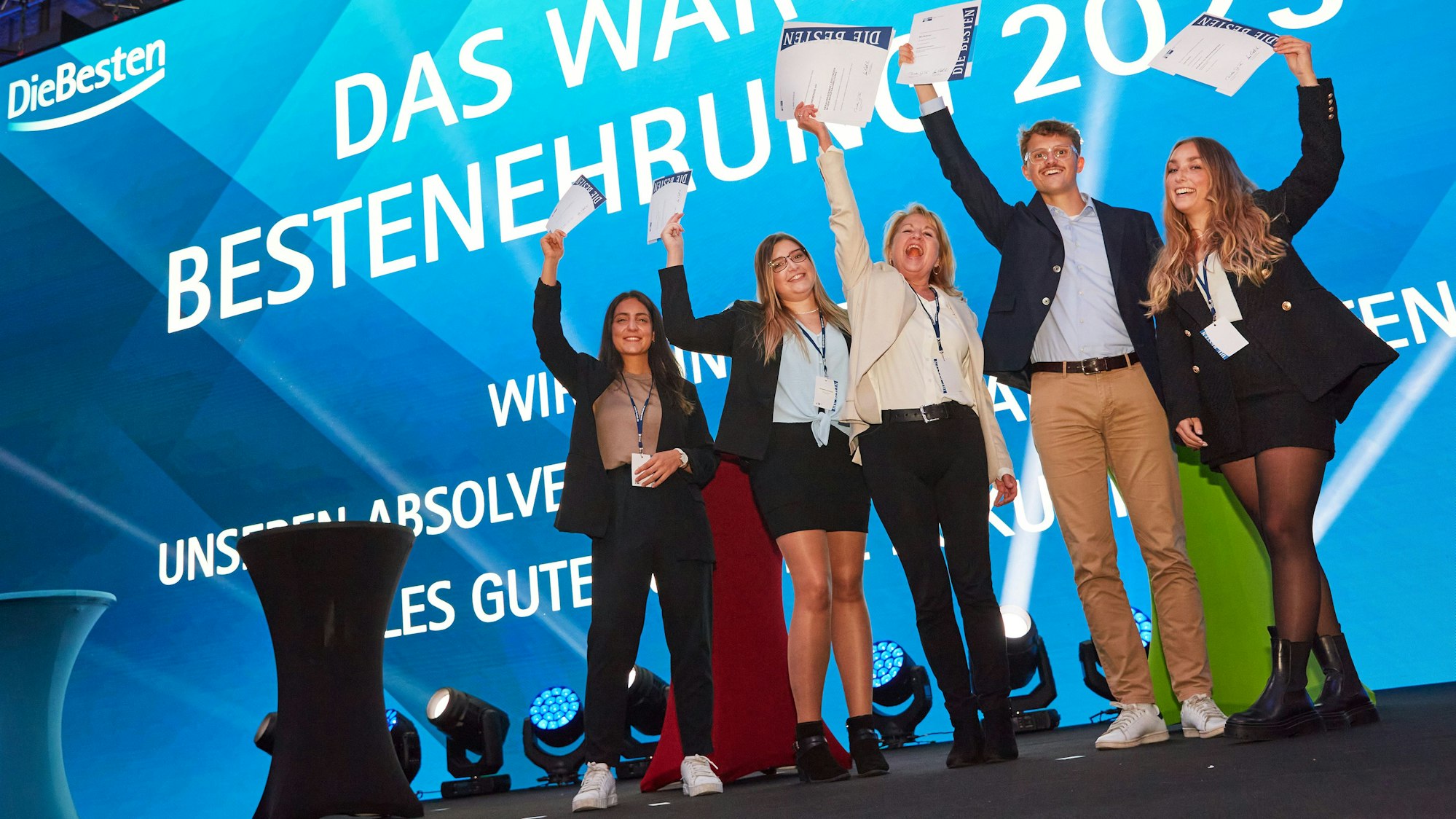 Ausgezeichnete Auszubildende halten auf einer Bühne lachend ihre Zertifikate in die Höhe.