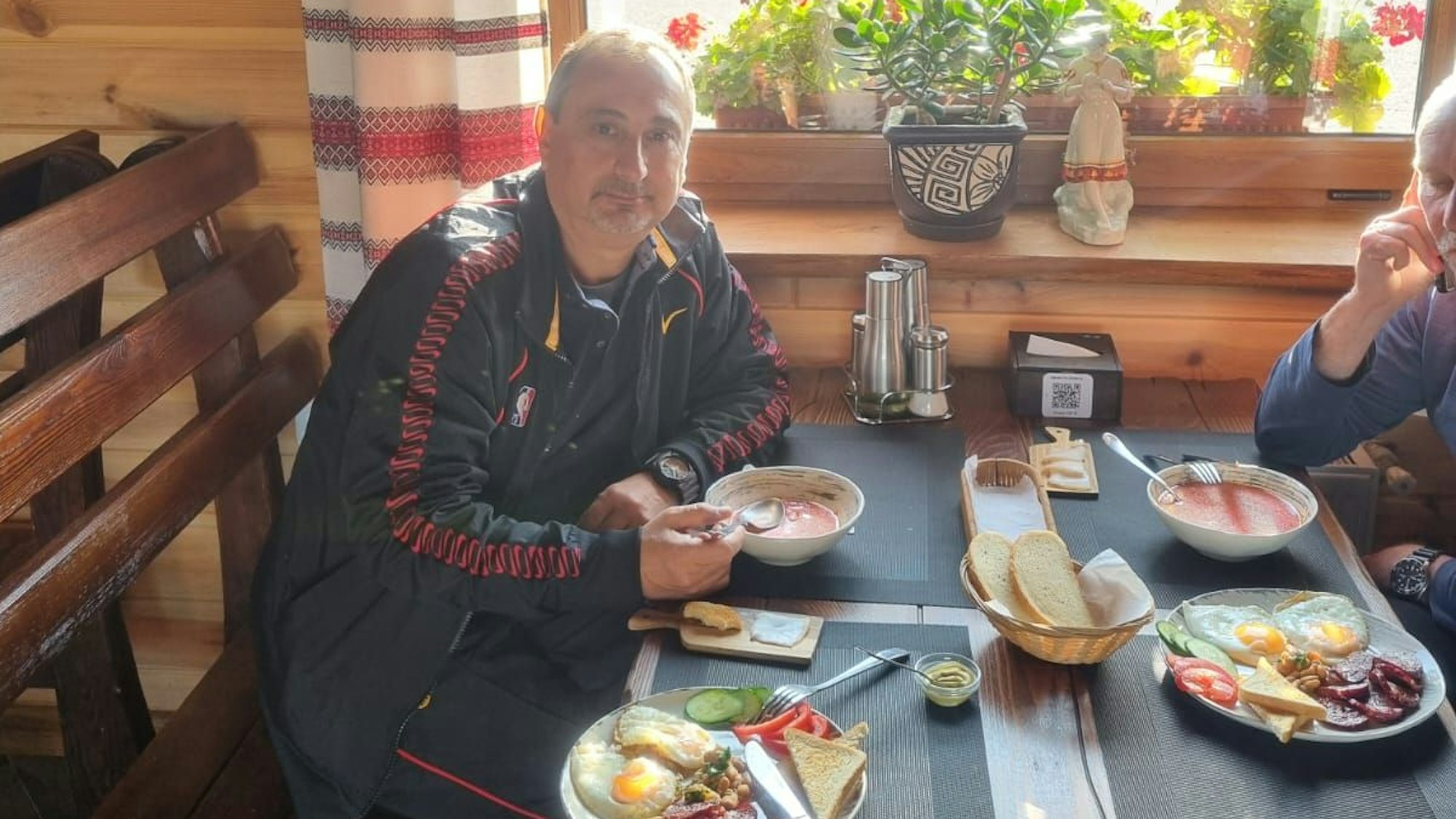 Ein Mann sitzt an einem Tisch und isst eine Suppe. Daneben steht ein Teller mit Gemüse, Aufschnitt und Spiegeleiern.