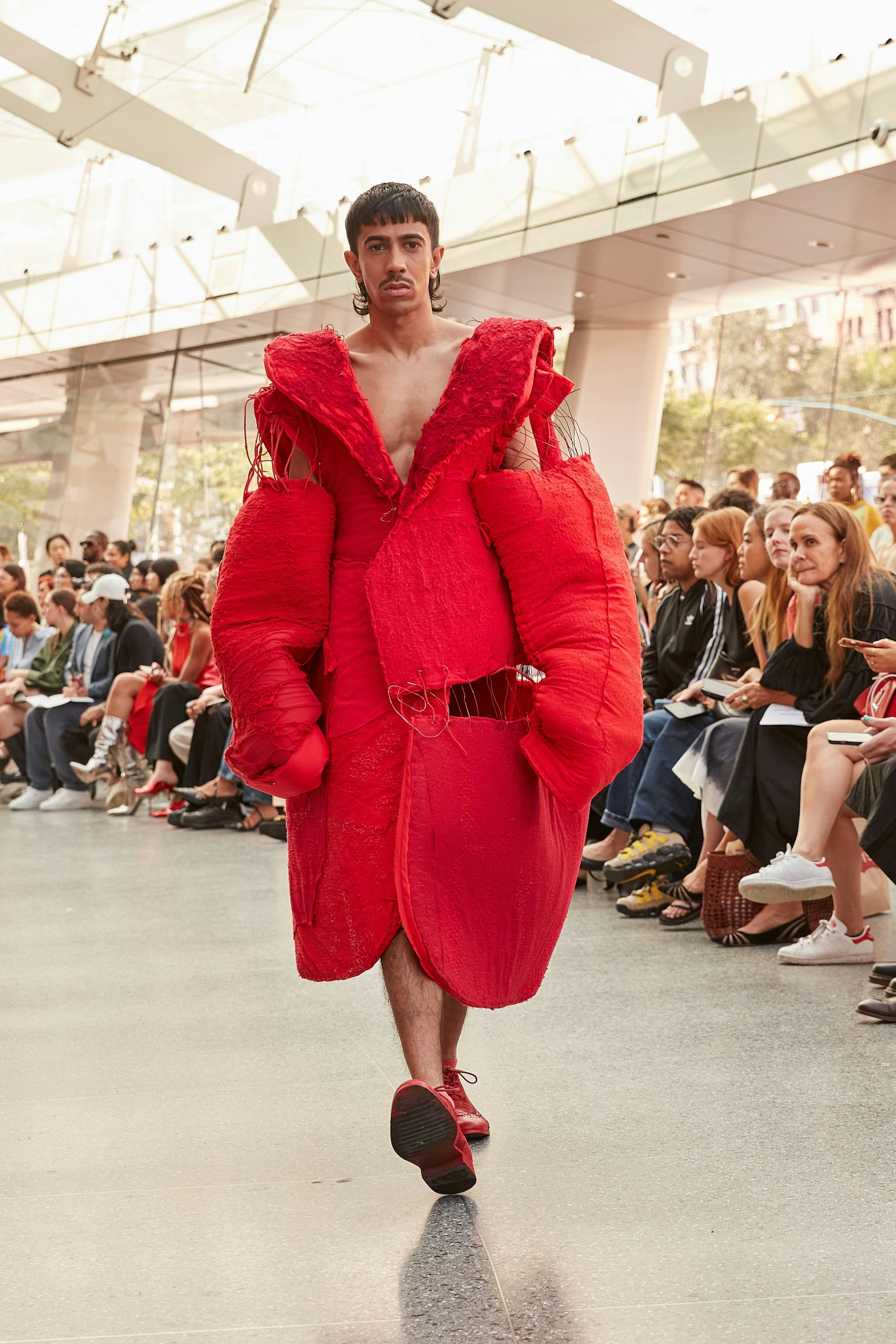 Ein männliches Modell trägt ein extravagantes rotes Kleidungsstück auf einem Laufsteg.