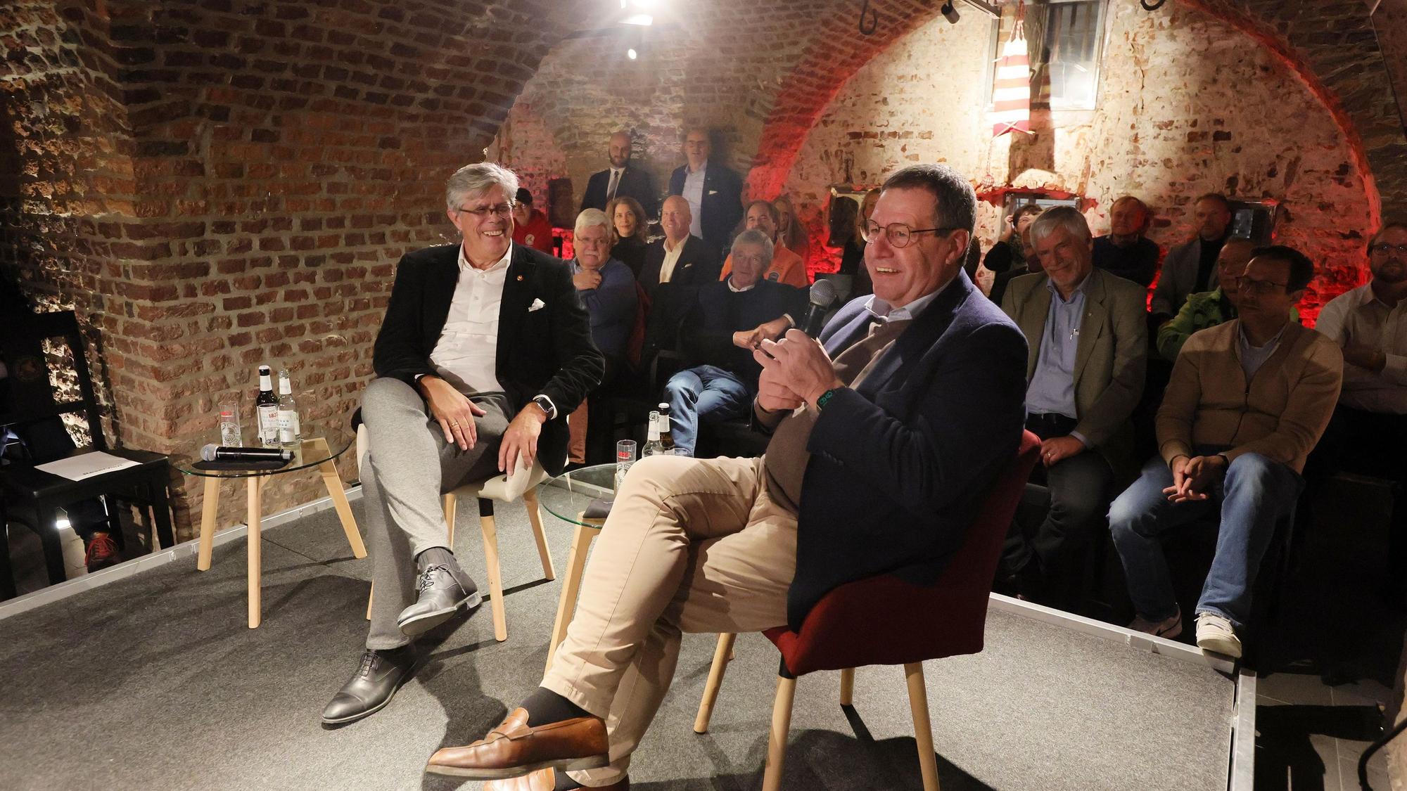 Zwei Männer sitzen auf einer Bühne, im Hintergrund sieht man ein Publikum.
