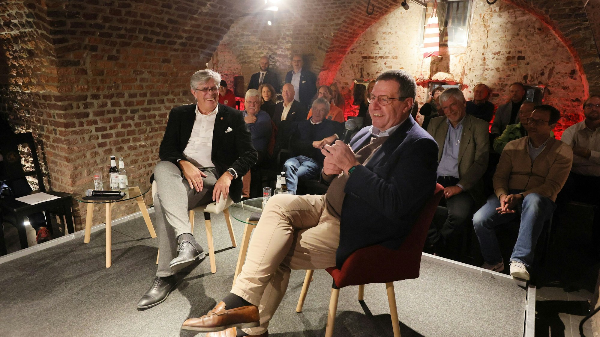 Zwei Männer sitzen auf einer Bühne, im Hintergrund sieht man ein Publikum.