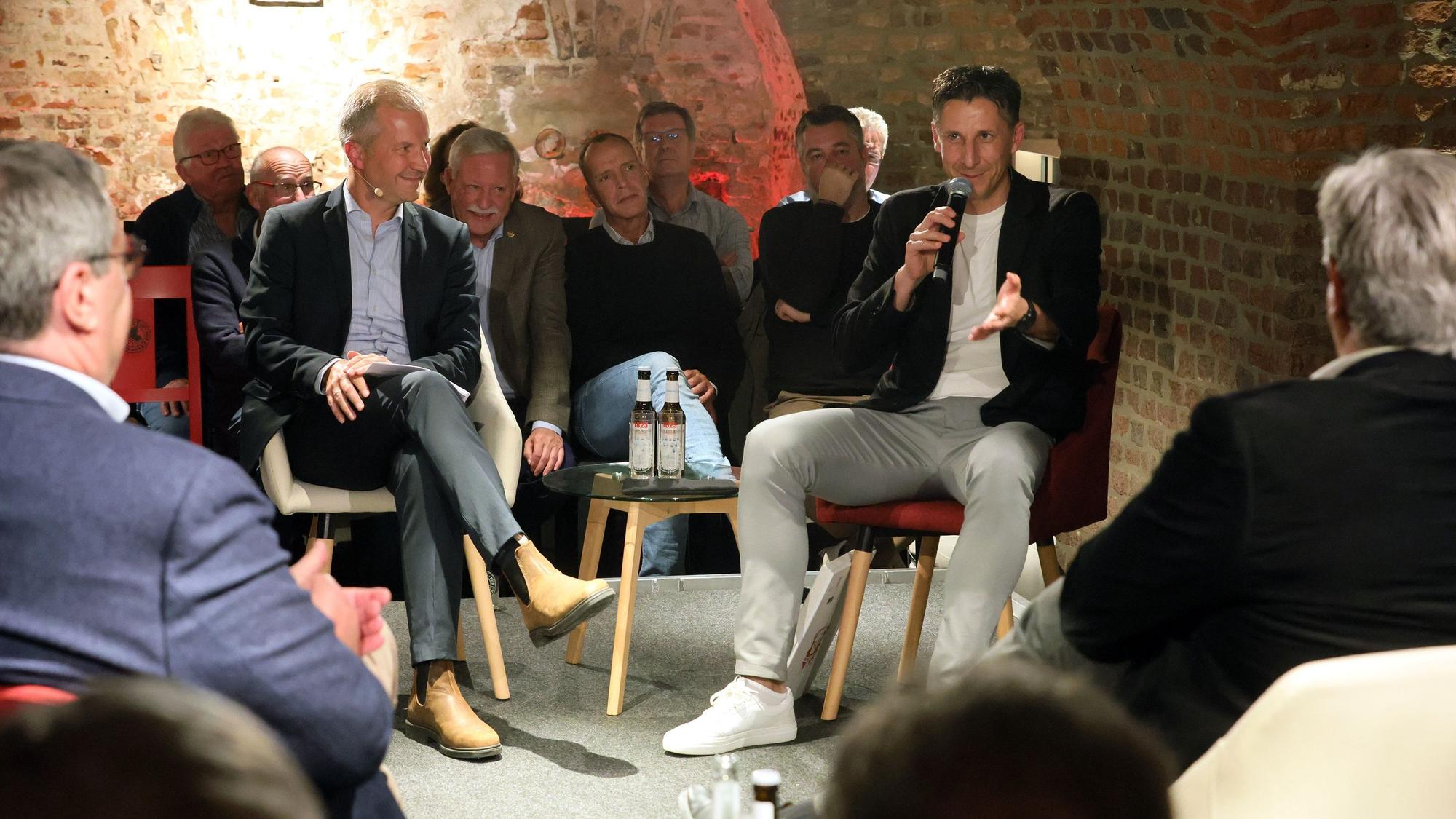 Vier Männer sitzen auf einer Bühne – im Hintergrund ist Publikum zu sehen.