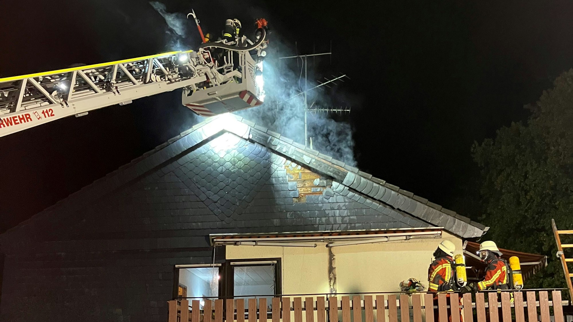Aus einem Dach steigt Qualm auf. Einsatzkräfte der Feuerwehr nähern sich dem Dach mit einer Drehleiter.