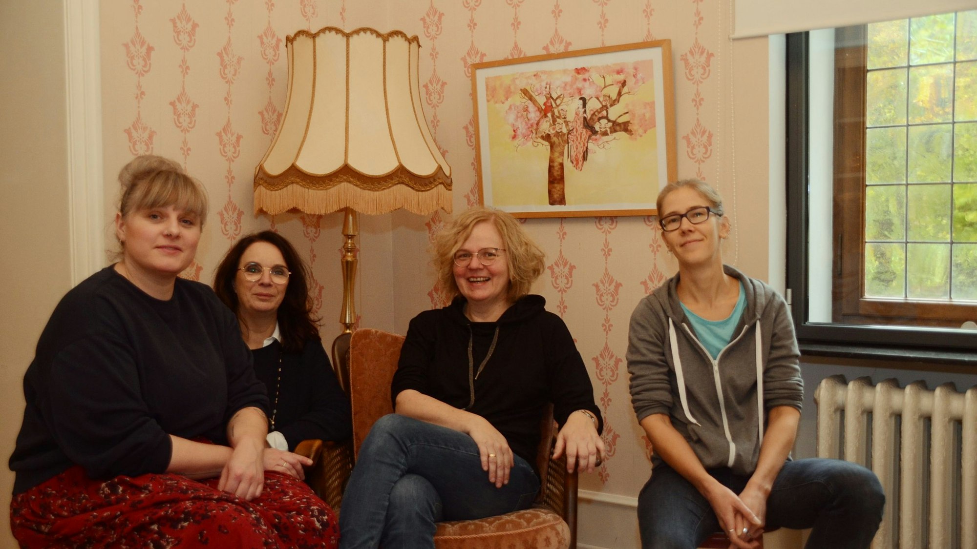 Vier Frauen sitzen vor einer gemusterten Tapete in hellrosa; hinter ihnen steht eine altmodische Stehlampe, an der Wand hängt ein Bild.