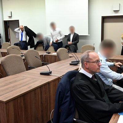 Zu sehen ist das Innere eines Gerichtsgebäudes in Köln. Die Angeklagten warten mit ihren Rechtsanwälten auf den Beginn des Prozesses.