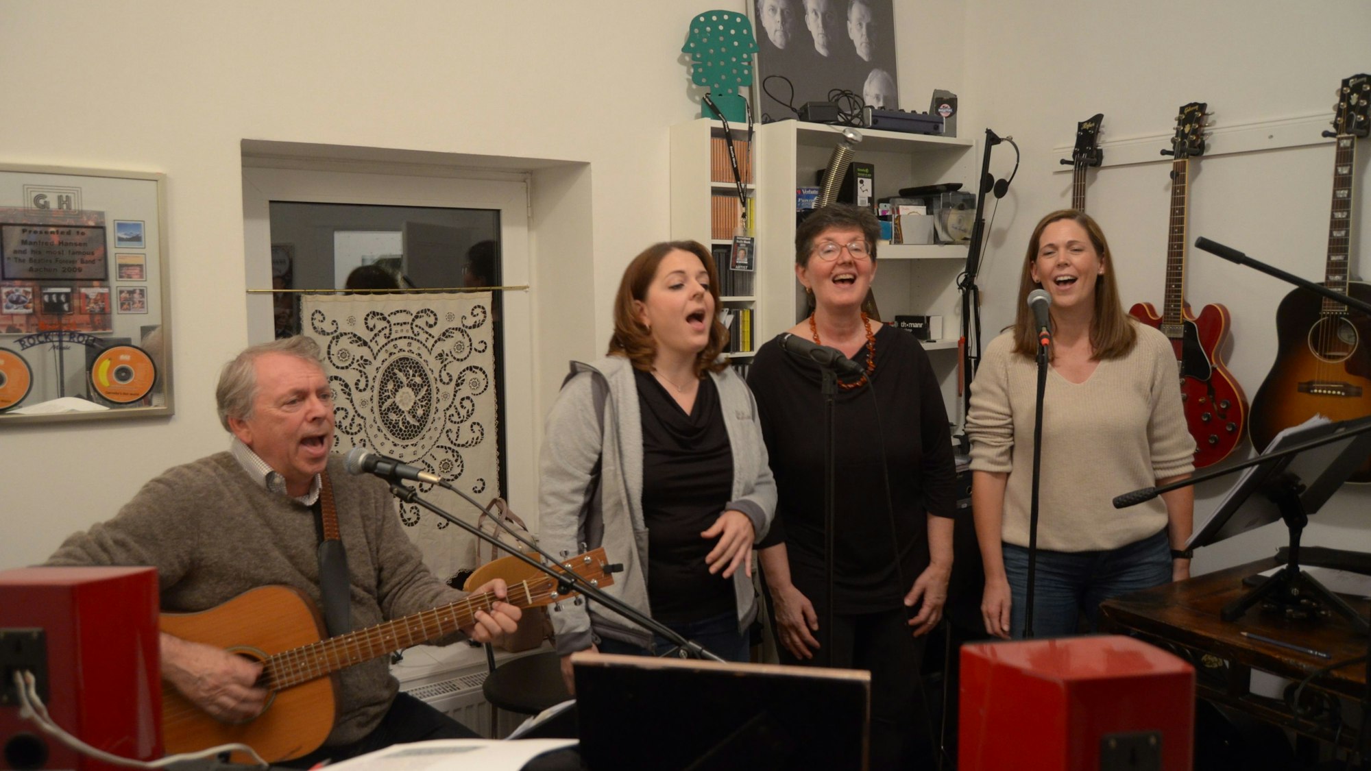 Vier Mitglieder der Band Lenny Pane proben ihr Programm mit Beatles-Liedern. Der Sänger und Gitarrist Manfred Hansen sitzt an einem Mikrofon, neben ihm stehen die Sängerinnen Franziska Lörch, Elise Kushner und Claudia Milz. Im Hintergrund hängen Gitarren und Plakate an der Wand.