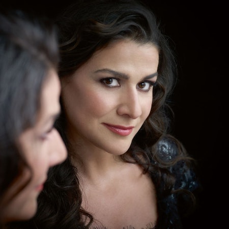 Eine Nahaufnahme von Cecilia Bartoli. Sie hat dunkle, gewellte Haare und braune Augen. Sie trägt Lippenstift und ein schwarzes Kleid.