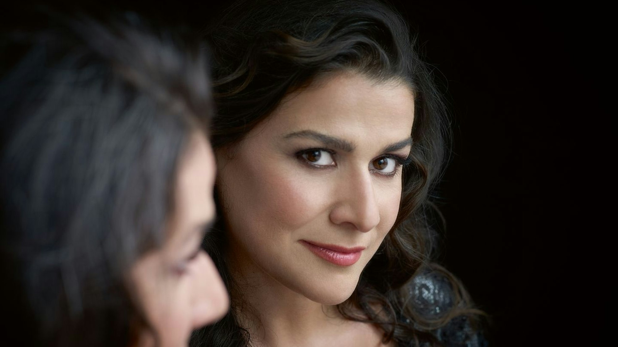 Eine Nahaufnahme von Cecilia Bartoli. Sie hat dunkle, gewellte Haare und braune Augen. Sie trägt Lippenstift und ein schwarzes Kleid.