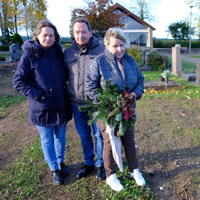 Gabi Große, Bernhard Hoffmann und Anja Hoffmann stehen auf dem Friedhof in Kommern an der Stelle, wo sich bis vor Kurzem noch das Grab ihrer Schwester Sabine befand. Gabi Große hält ein Grabgesteck in der Hand.