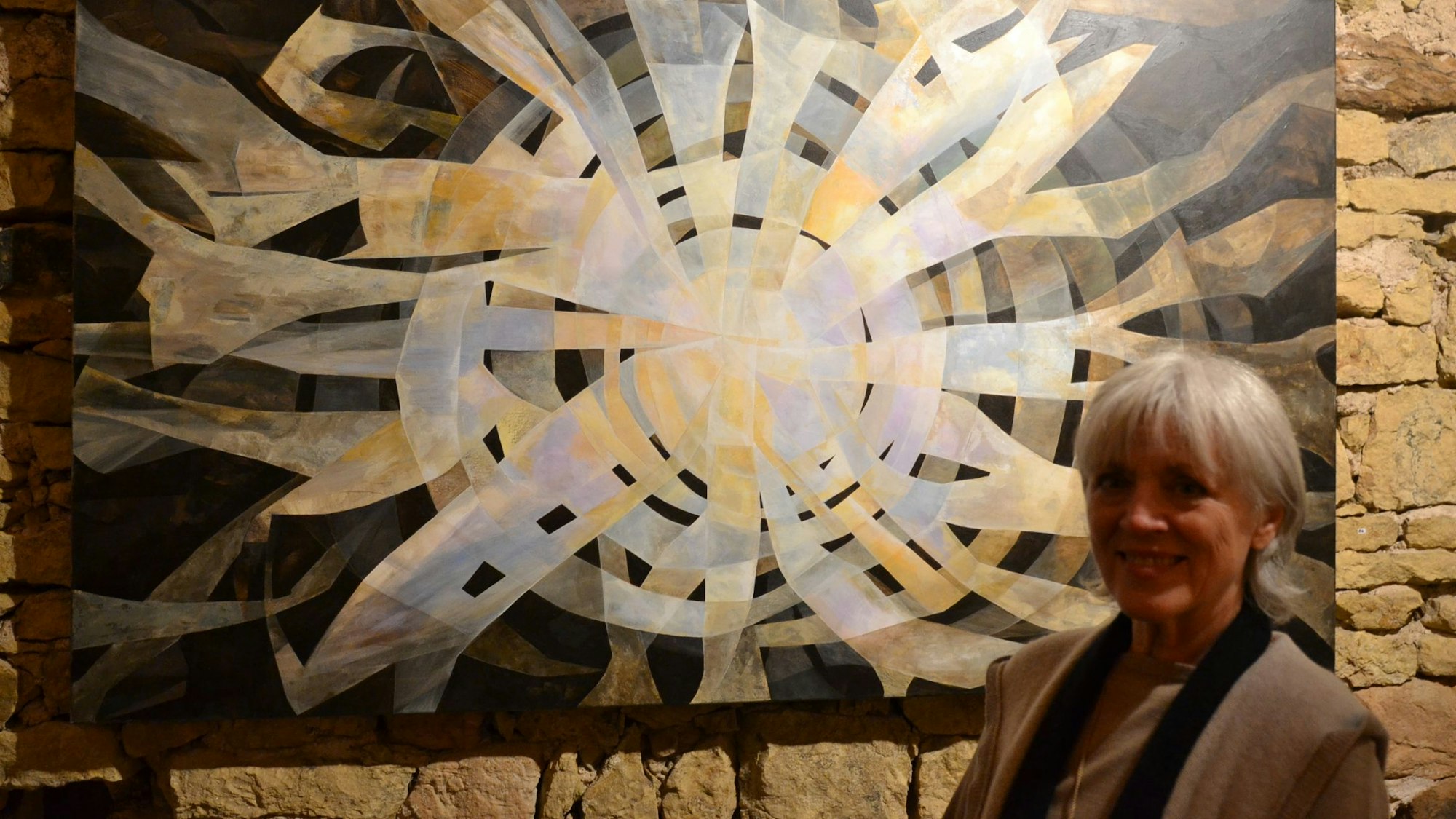 Gabriele Latzke steht vor einem Gemälde. Helle Streifen fließen von der Mitte des Bildes nach außen, der Hintergrund ist schwarz.