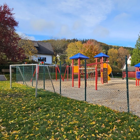 Zu sehen ist das Tor des Bolzplatzes und mehrere bunte Geräte des Spielplatzes.