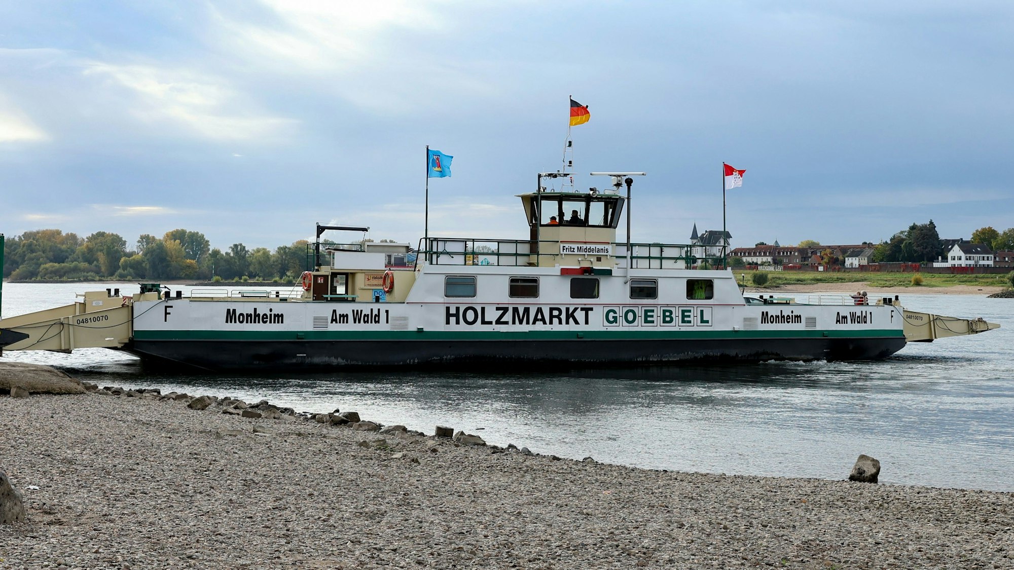 Die Rheinfähre „Fritz Middelanis“ verkehrte zwischen Köln-Langel und Leverkusen-Hitdorf. Die Fähre, die auch Autos transportierte, liegt am linken Rheinufer. Im Vordergrund ist Uferstrand zu sehen, im Hintergrund das gegenüberliegende bewaldete Rheinufer bei Hitdorf.