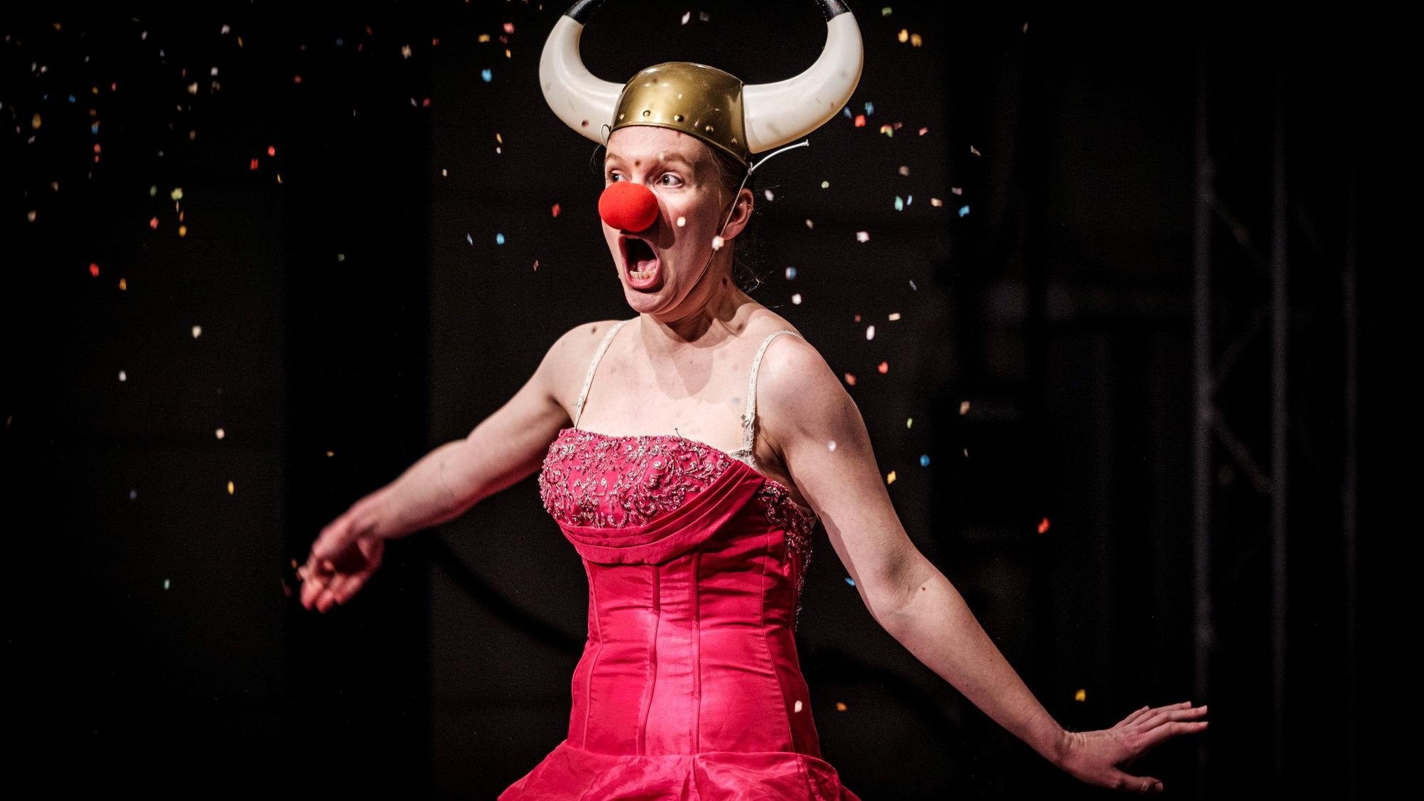 Eine Schauspielerin im roten, schulterfreien Kleid trägt eine rote Clownsnase und einen gehörnten Wikingerhelm, Szene aus „Frühlingserwachen“ im Theater der Keller