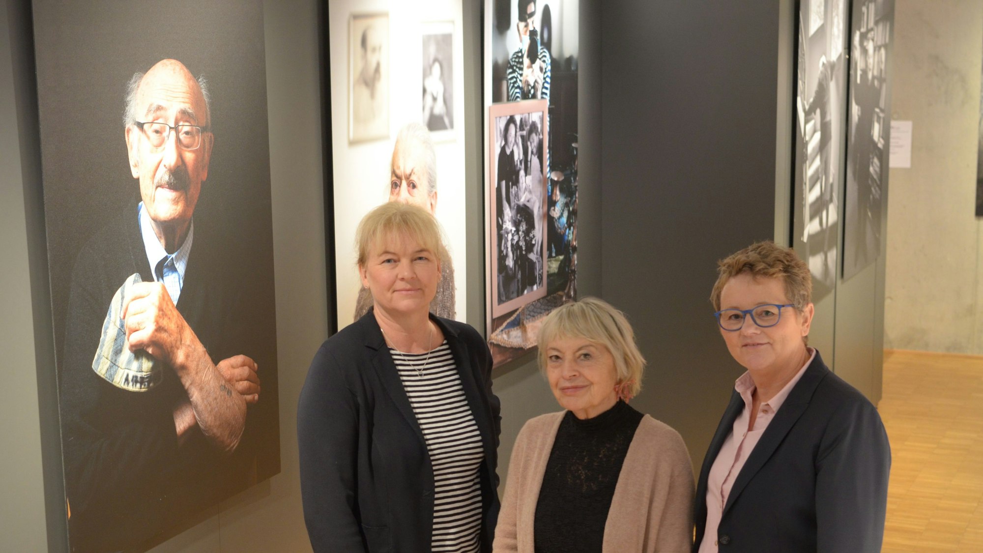 Dr. Heike Lützenkirchen (l.) und Petra Goerge (r.) stellen im Stadtmuseum Euskirchen mit Kuratorin Gisela Kayser die Sonderausstellung „Lonka Projekt“ vor. Sie stehen vor einer Reihe von Porträts von Holocaust-Überlebenden.