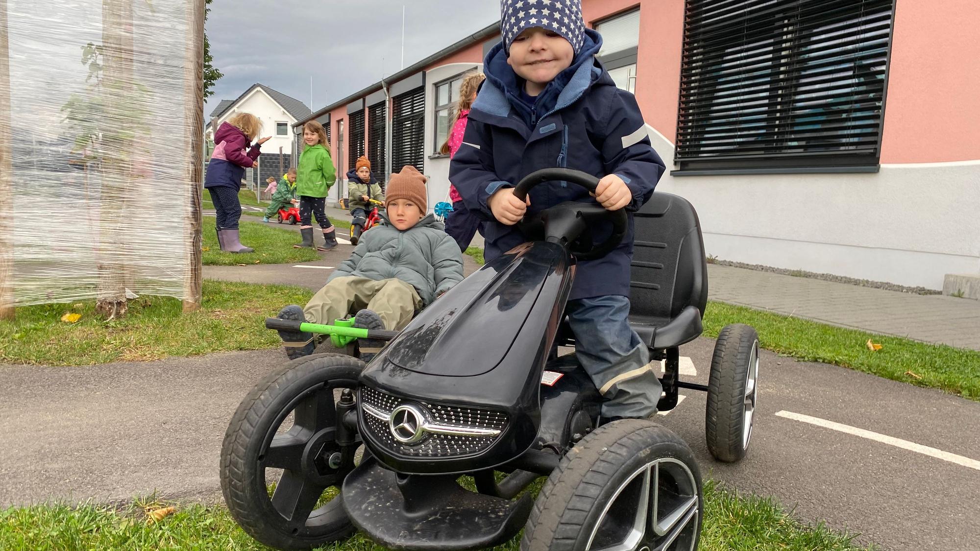Ein Junge fährt auf einem Kettcar. Es hat eine Kühlerhaube wie ein Mercedes und dicke Reifen.
