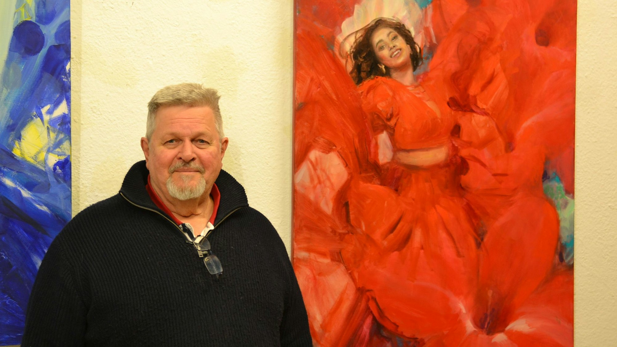 Galerist Rüdiger Axel Westphal steht vor einem Gemälde von Olga Müller, das eine Salsa-Tänzerin zeigt, deren rotes Kleid in ein Meer aus roten Blüten übergeht.