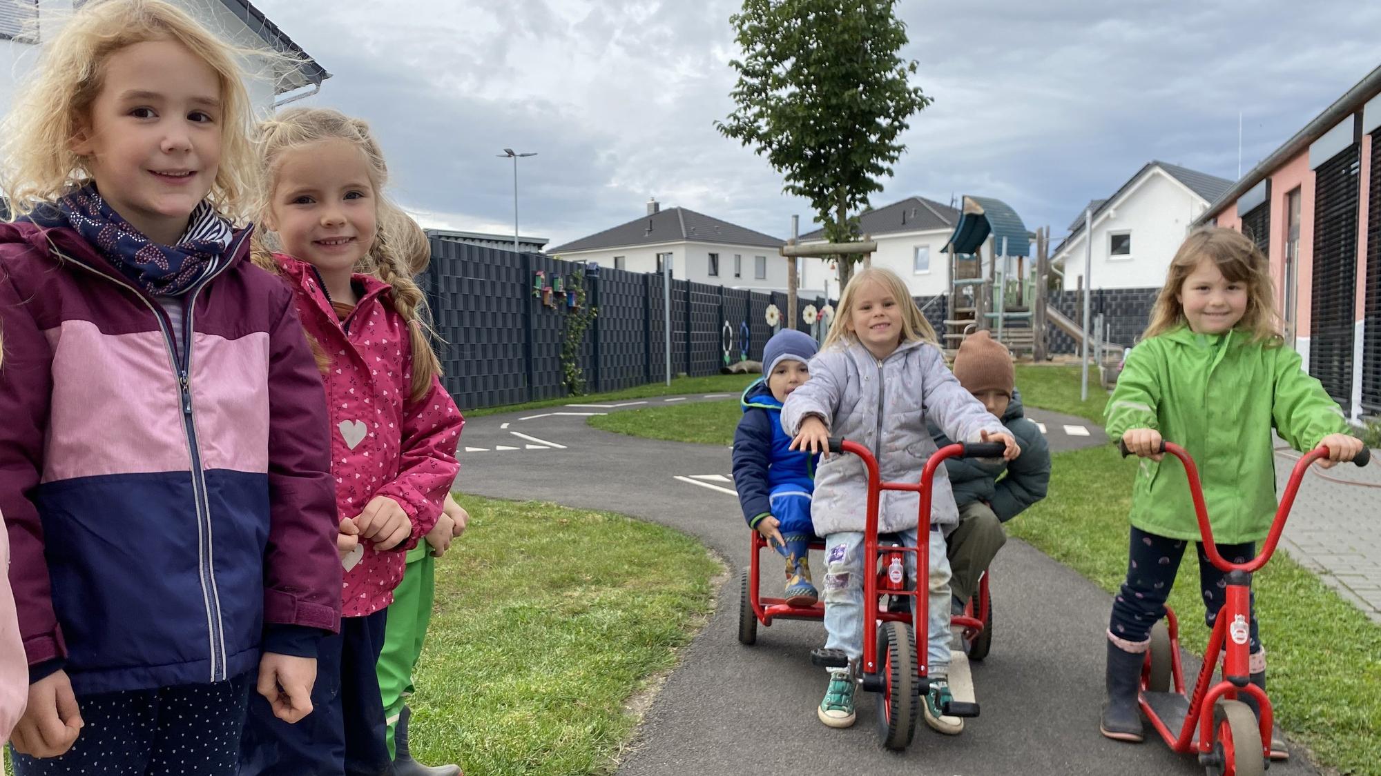 Zwei Mädchen stehen am Straßenrand. Eine Dreiradfahrerin und eine Rollerfahrerin halten auf der schmalen Straße.