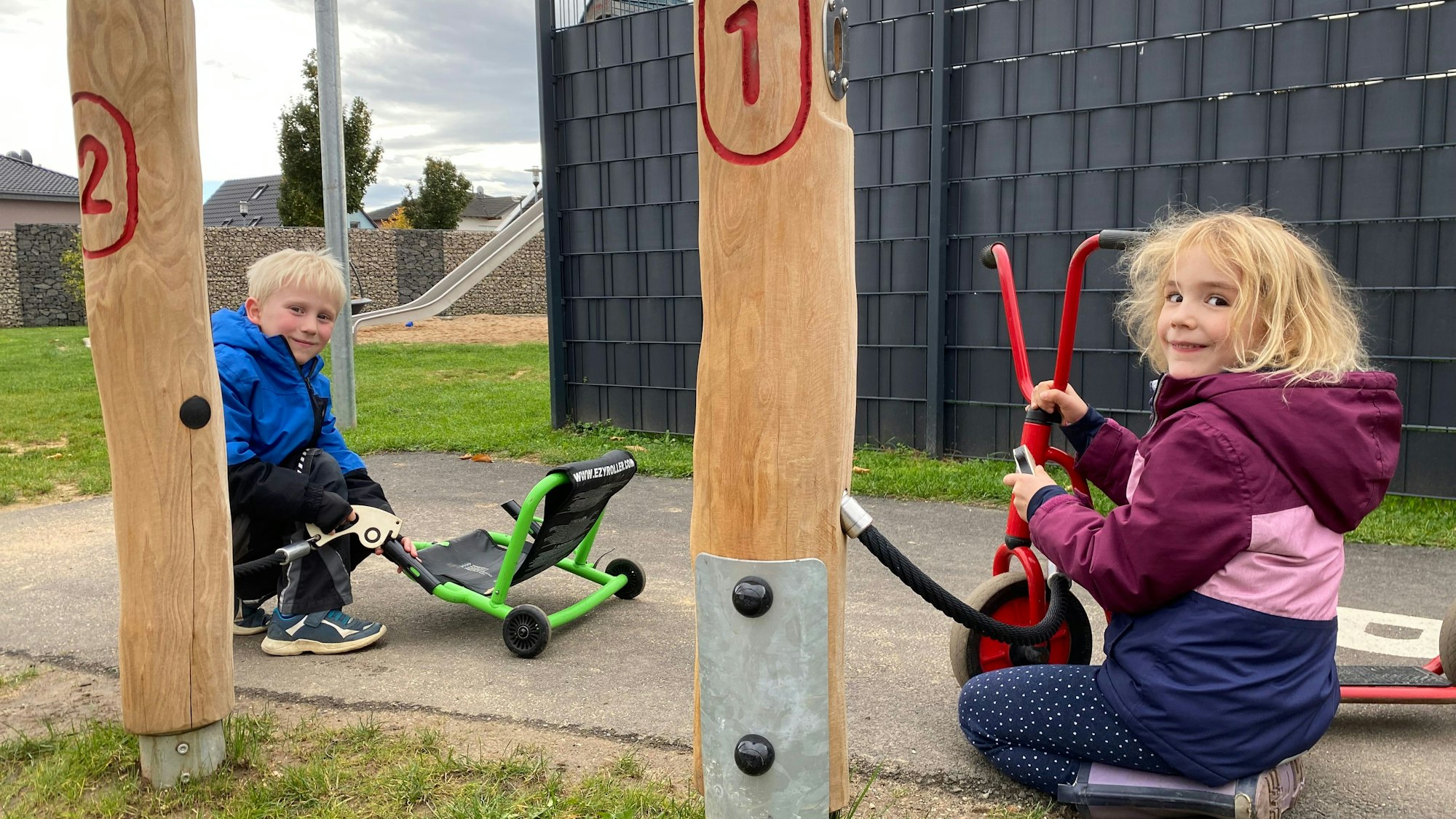 Zwein Kinder tun so, als würden sie ihre Kettcars betanken. Die Zapfsäulen sind zwei nummerierte Holzpfahle.