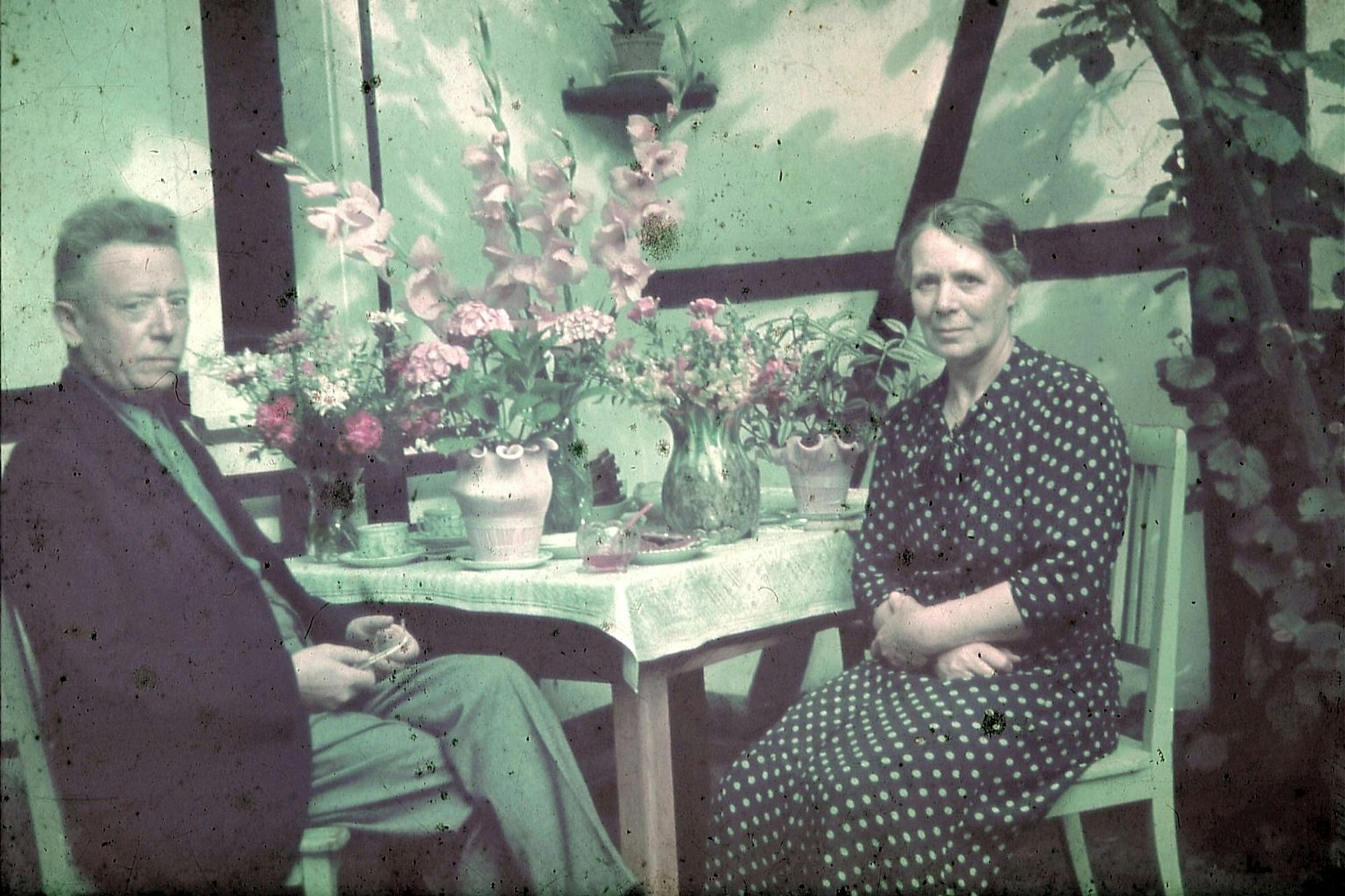 Mann und Frau sind auf diesem verblassten Farbbild an einem gedeckten Tisch vor einer Fachwerkwand zu sehen.