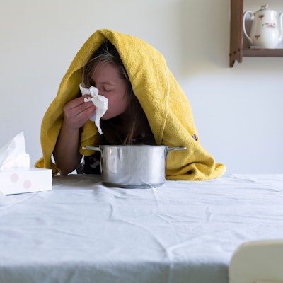 Frau mit Handtuch über dem Kopf sitzt vor einem Kochtopf am Tisch und putzt sich die Nase.